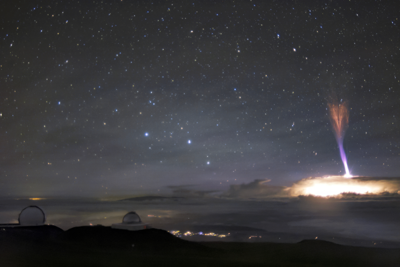 This image shows a wide view of a horizon, with the distinctive round top of an observatory on top of a dark mountain on the left and a big storm cloud over the horizon on the right, with a wide view of distant stars clear in the night sky above. The storm cloud is lit from within by lighting, with a white-to-purple to red finger of light extending from the top of the cloud up. The top of this finger of light extends much higher in the sky than the distance from cloud-to-ground. At the top of this jet, the light is red and spreads in smaller fingers, still pointing up, like a tight bouquet of grass blades or a quiver of arrows. 