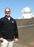Portrait photo of a man standing outside with a large telescope in the background.