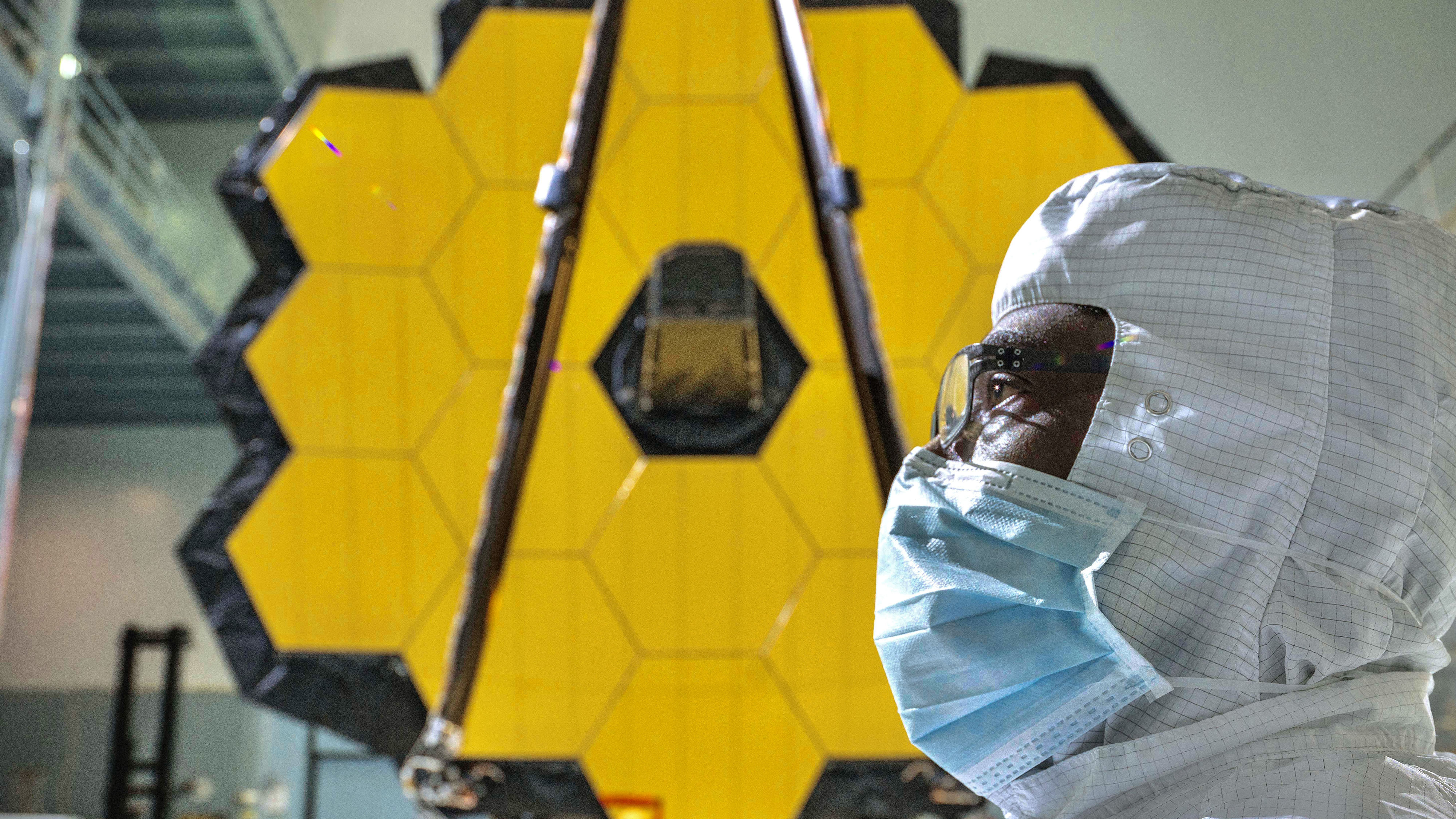 In the foreground, the side profile of Webb lead photographer Chris Gunn wearing glasses and a mask in a cleanroom suit stares intently off screen to the left. Directly behind, the slightly blurry, full honeycomb like golden mirror of the James Webb Space Telescope is in clear view taking up most of the frame.