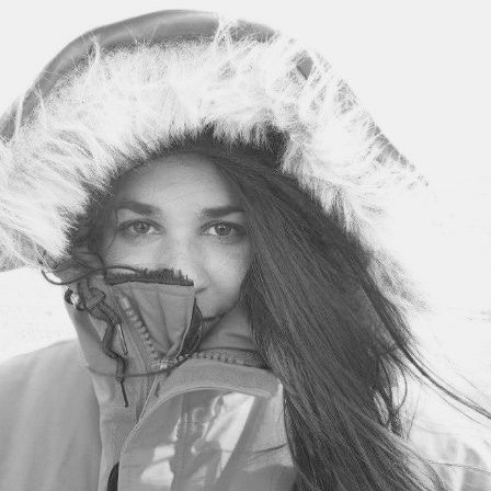 Black and white photo of Kelsey Bisson with long dark hair in a winter jacket. The jacket is covering her mouth and nose.