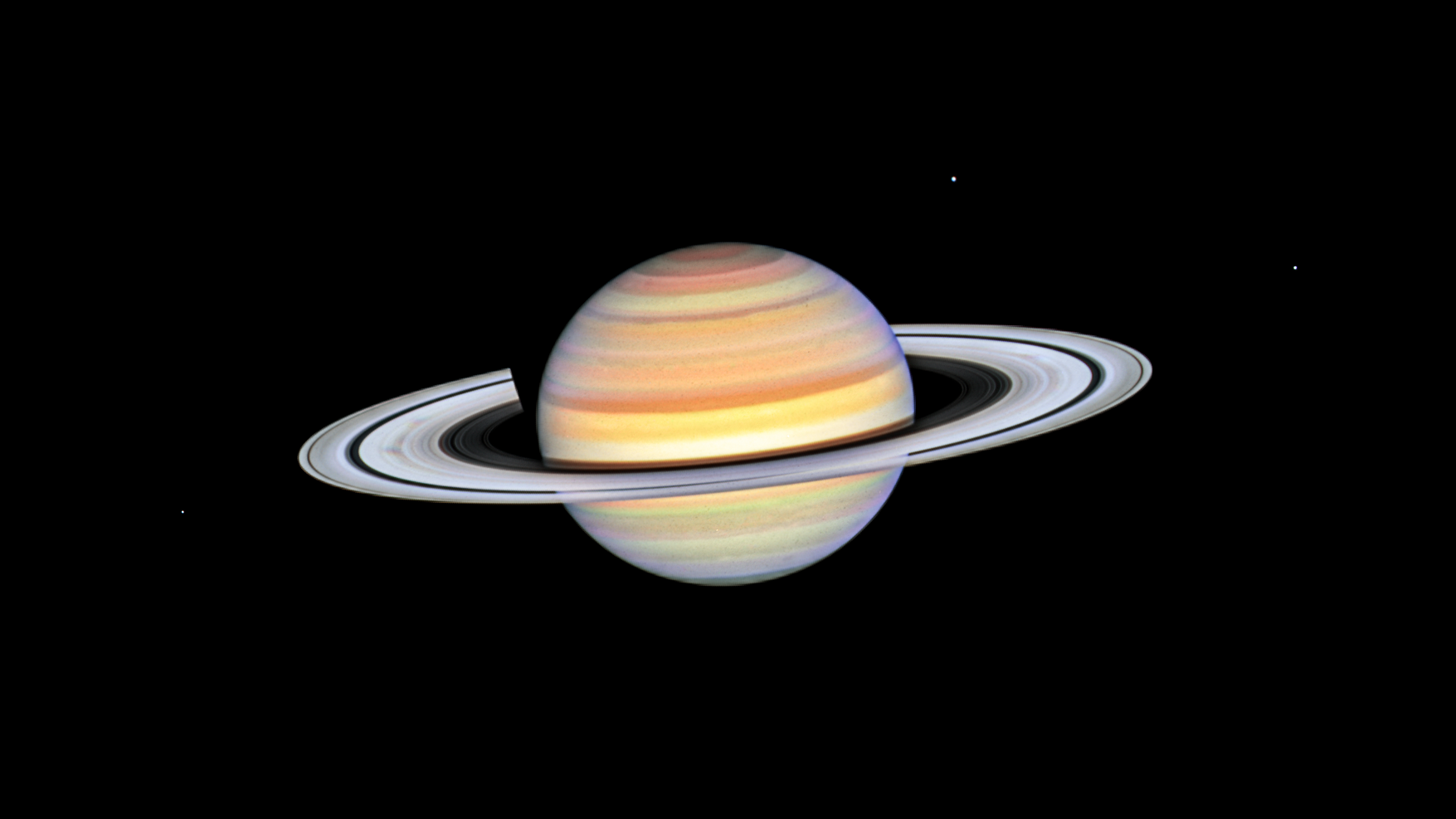 Colorful stripes in yellow, white, reddish-orange, pink, and green cover the planet. Saturn is tilted slightly toward us allowing the Sun to illuminate the top of its rings. The planet's shadow is cast toward the back and left of the planet. Saturn's moons Dione, and Enceladus are visible to its upper right, while its moon Mimas is just below and to the left of the planet's rings.