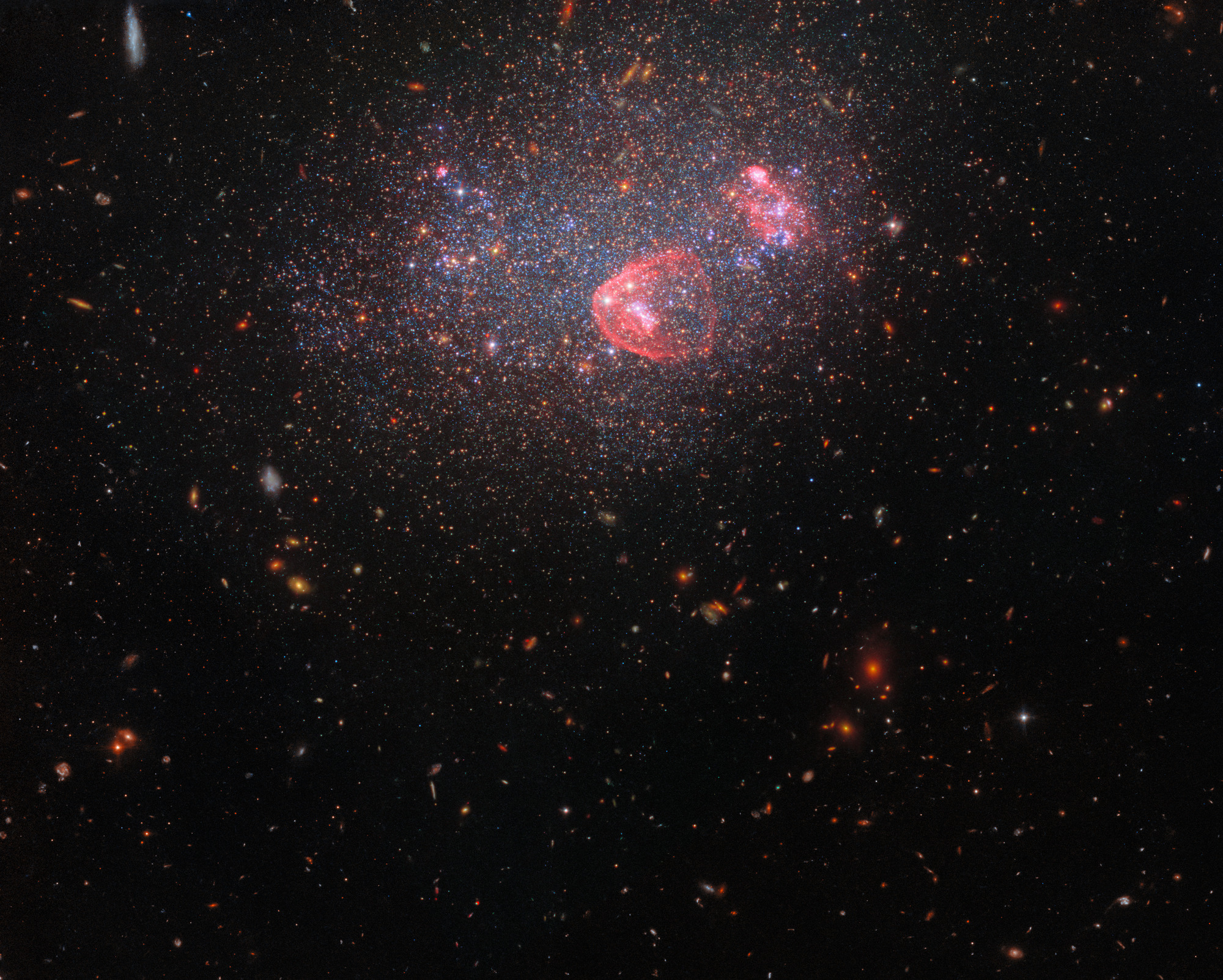 A field of galaxies. UGC 8091 is at the top and center of the image. It appears as a haze of stars through which more distant galaxies are visible. Two, bright, pinkish-red nebulae are also visible.