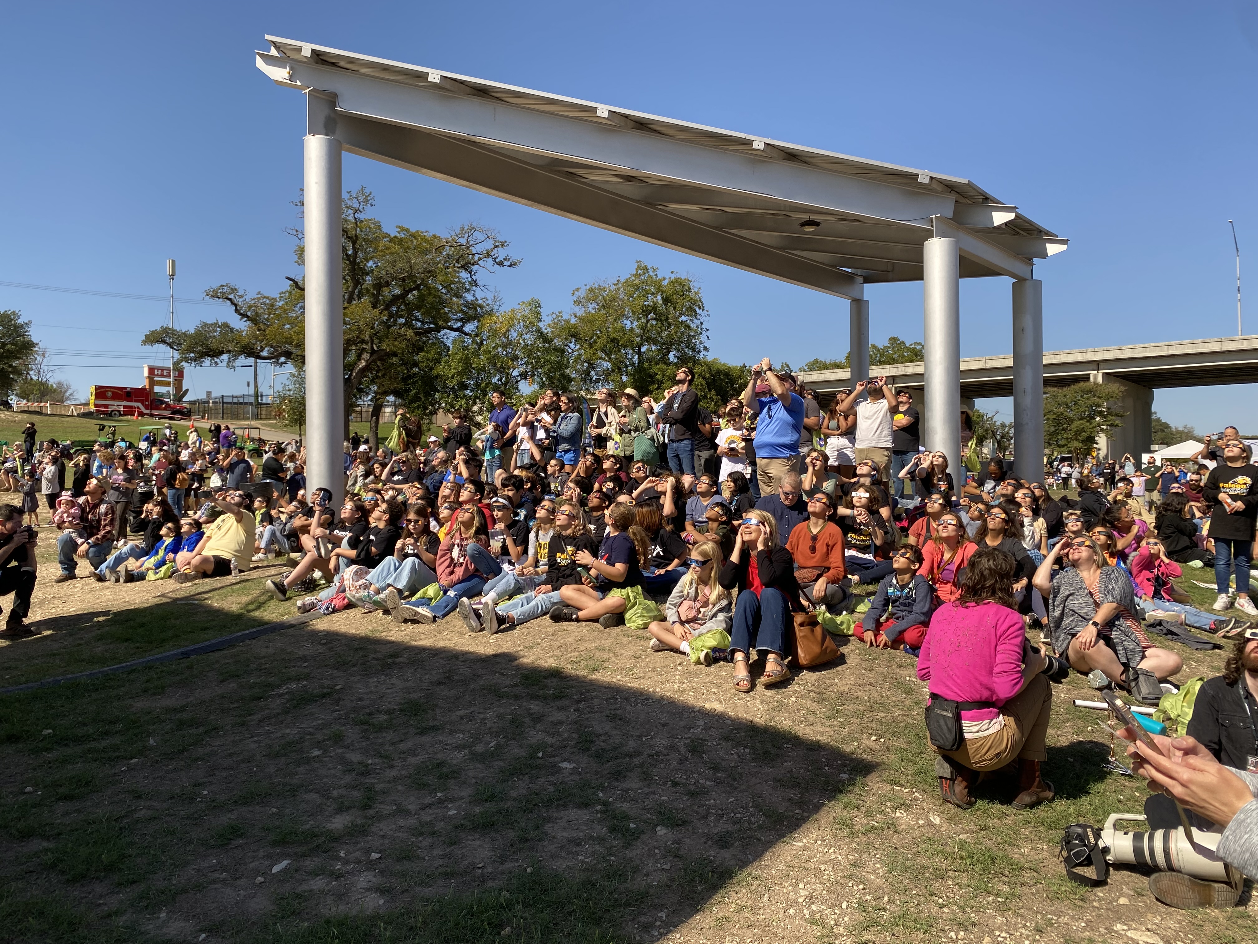 A large group of people sit on grass below a pavilion, holding eclipse glasses up to their eyes.
