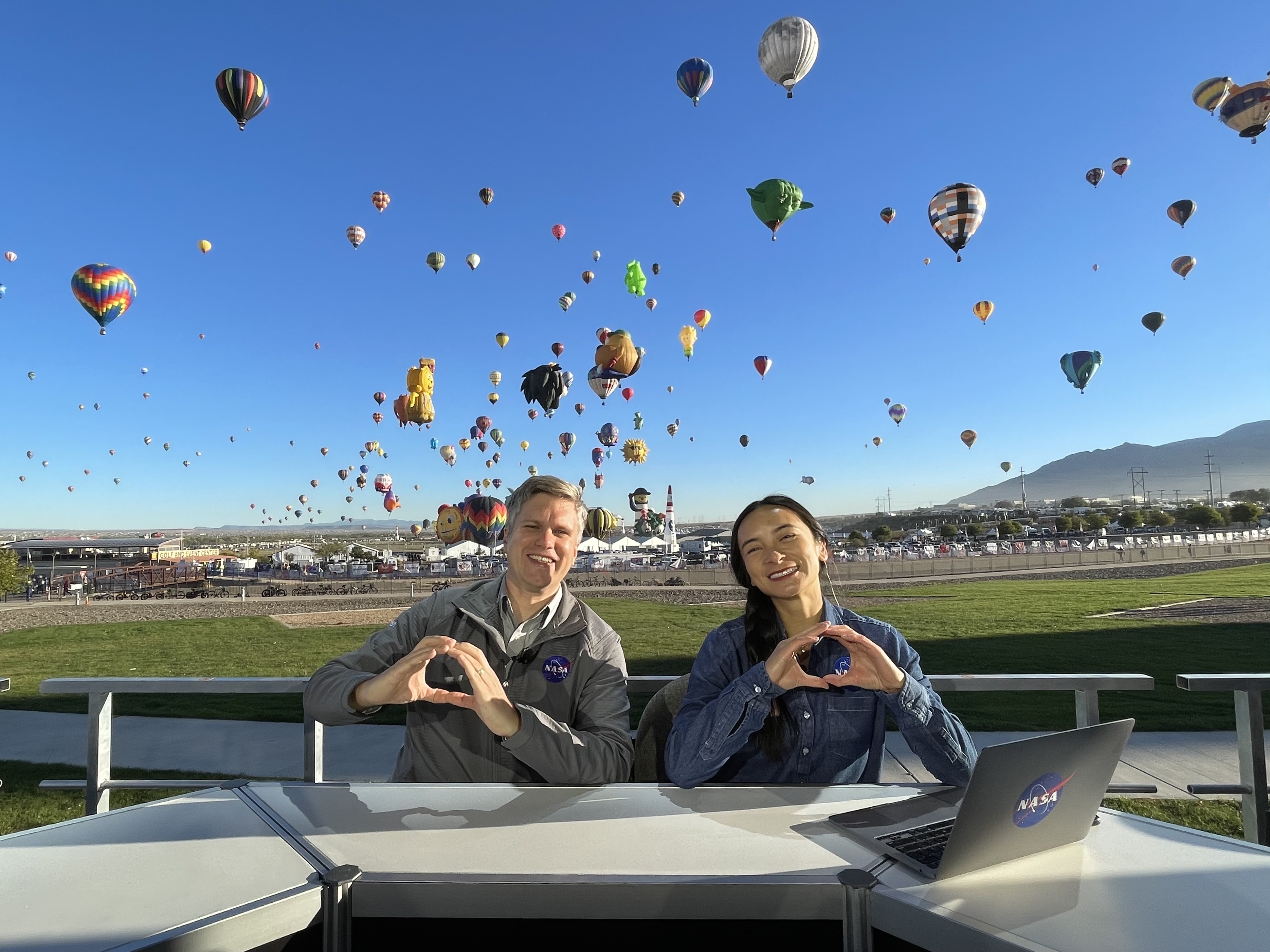 A man and a woman sit at a desk, making hearts with their hands. Behind them is a large green field, with many hot air balloons on the field and in the sky