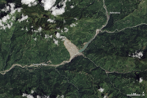 A true-color satellite image shows a green, mountainous area bisected from top right to bottom left by a large river. Clouds float over the peaks. A large brown-gray triangular scar starts at the top of a mountain and widens down to and across the riverbed. The river has cut through this material and washed it downstream, as evident by the brown color in the riverbed, which contrasts with the green color of the river upstream of the slide. 