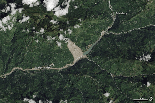 A true-color satellite image shows a green, mountainous area bisected from top right to bottom left by a large river. Clouds float over the peaks. A large brown-gray triangular scar starts at the top of a mountain and widens down to and across the riverbed. The river has cut through this material and washed it downstream, as evident by the brown color in the riverbed, which contrasts with the green color of the river upstream of the slide. 