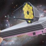 A hand holding a paper model of the James Webb Space Telescope on a cosmic background of stars and a cloudy galaxy.