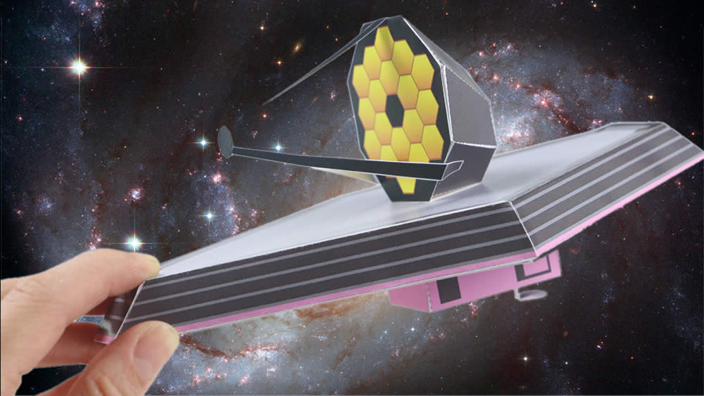 A hand holding a paper model of the James Webb Space Telescope on a cosmic background of stars and a cloudy galaxy.