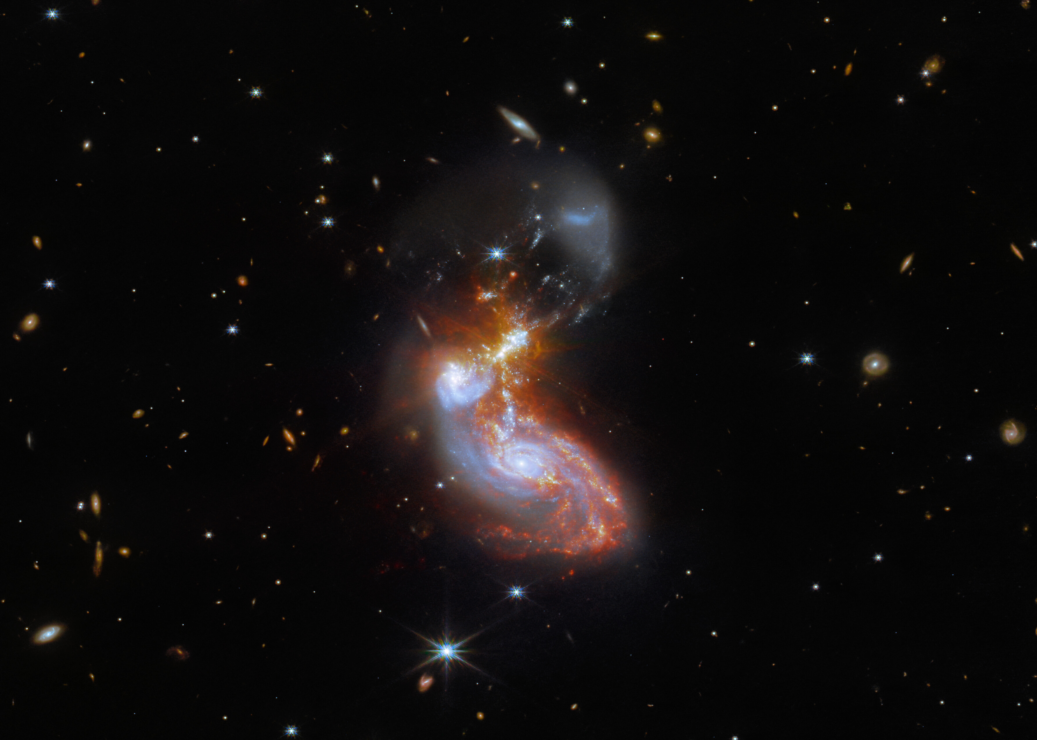 Together, two merging galaxies form a bean-like shape just below the center of this image. The cores of the galaxies are colored blue, and one is above and to the left of the other. These two cores are surrounded by bright tendrils of coral star-forming regions, which stretch above and below. To the right of the top galactic core is a particularly bright star-formation clump with faint diffraction spikes. The lower galaxy is a mostly regular spiral shape, while the upper galaxy has been distorted into a much more condensed bun. There is also a semi-transparent arc of pale dust above both merging galaxies. The background is black, and many tiny distant galaxies dot the scene.