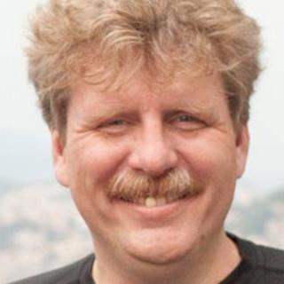 Portrait photo of a smiling man with curly blonde hair and a mustache.