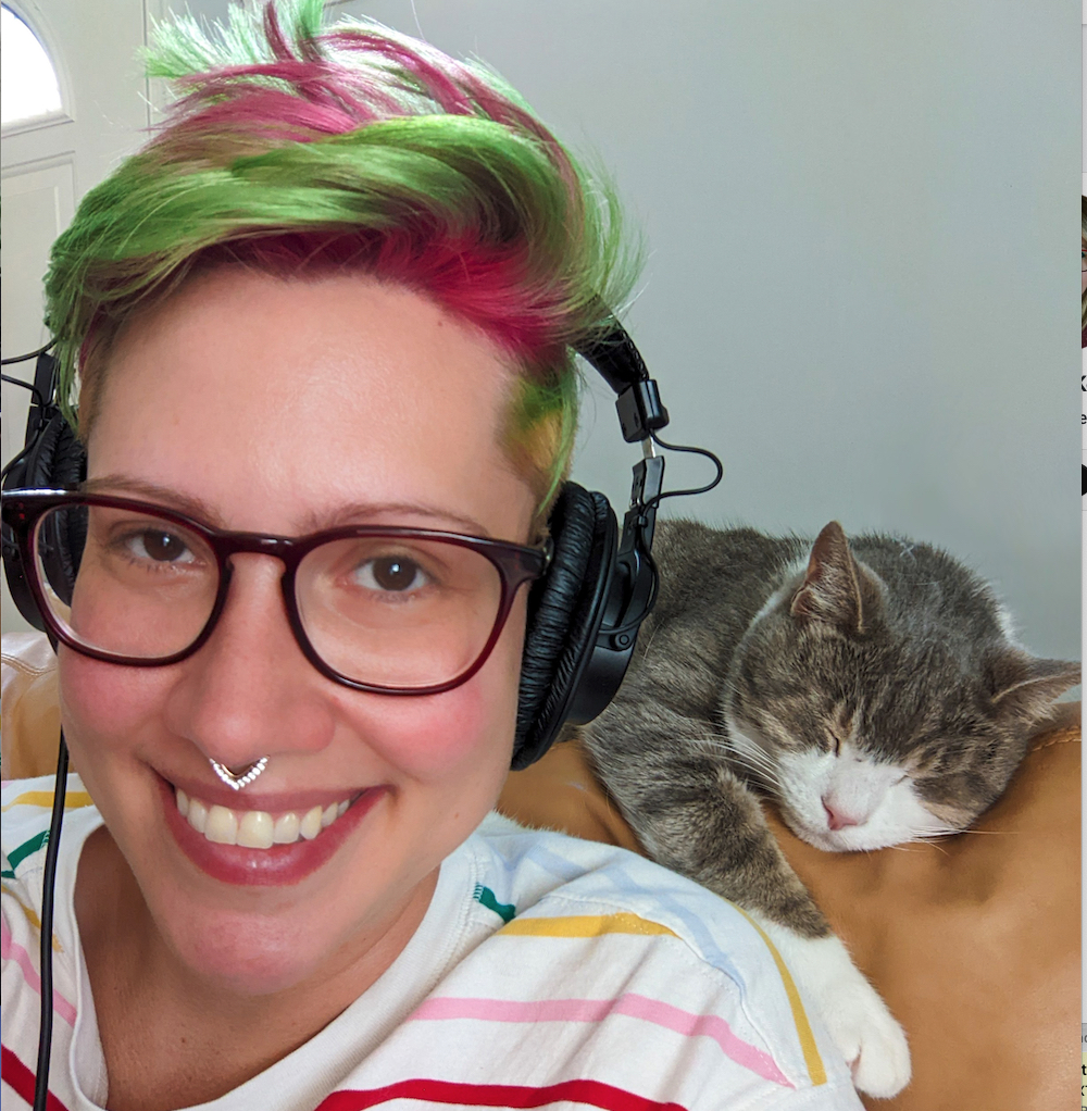 Portrait photo of a person wearing glasses with a nose piercing and hair died bright green and red. A cat sits above their shoulder on the back of the chair.