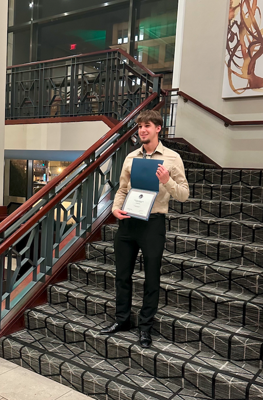 Leo Ravelo smiles and stands on a stairway, holding a certificate open for the photo as he accepts his scholarship at the 2023 ASGSR Conference in Washington, DC.