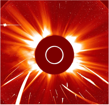Like all SOHO data, this image is rendered in reds. A dark red occulting disk in the center of the square image blocks the bright orb of the sun. A white circle with a diameter of one third of the occulting disk indicates the size of the Sun itself. We see the sun’s corona and solar outflows emanating from behind the disk, showing bright white adjacent to the circle and cooling to dark oranges at the edge of the image. Five fat white streaks and sixteen skinny streaks, all in the bottom half of the image, represent the tracks of sungrazing comets.