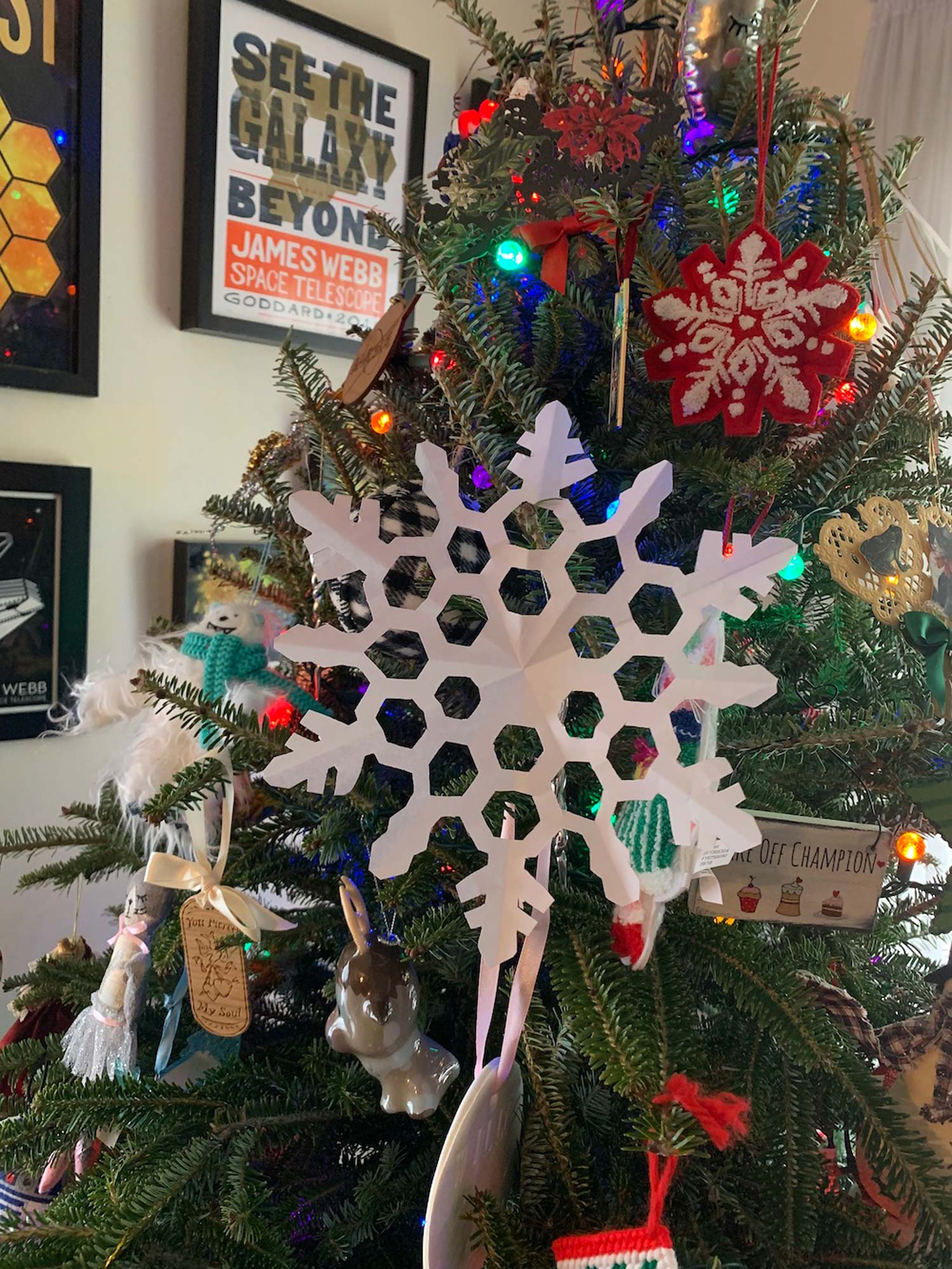 Christmas tree with lights and multiple ornaments including a paper snowflake ornament resembling the mirror pattern of the James Webb Space Telescope. On wall behind the tree hangs various frames with references to the James Webb Space Telescope.