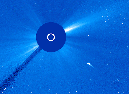 Typical of LASCO C3 data, the image is rendered in blues. A dark “occulting disk,” which is what we call the structure in the telescope that blocks the direct (blinding) sunlight, blocks the Sun. A white circle on the occulting disk indicates the size of the Sun itself. We can see the corona radiating outward in uneven streaks. Stars dot the field behind the sun. A white line representing a comet streaks through space just below and to the right of the Sun. As it reaches and passes behind the occulting disk, a white burst erupts from the other side.
