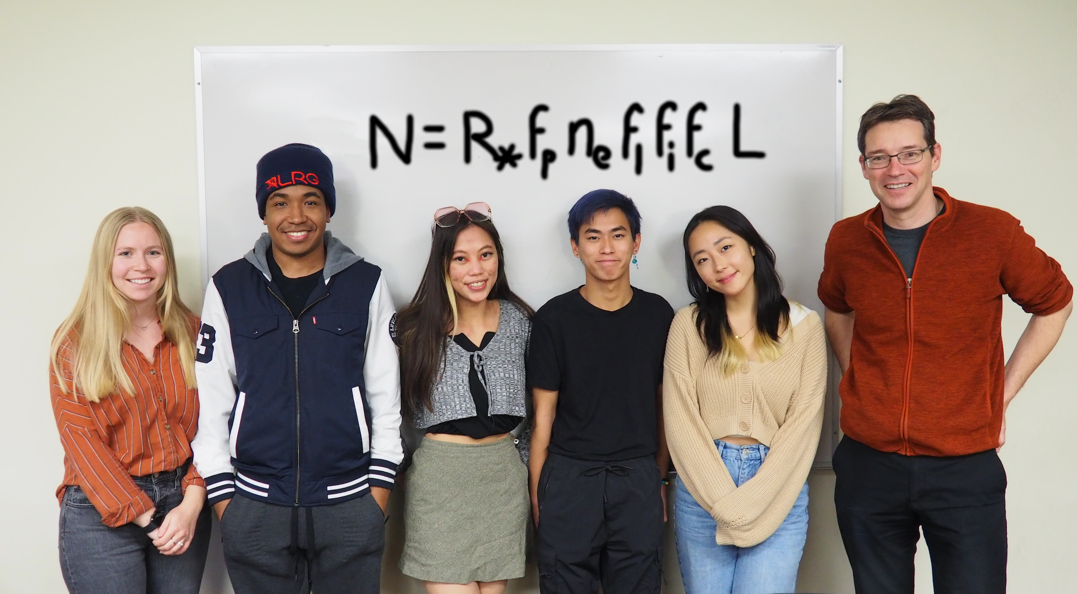 A group of 6 men and women are standing and smiling in front of a white board with the equation N-R*fpnef1fifcL written in large black print.