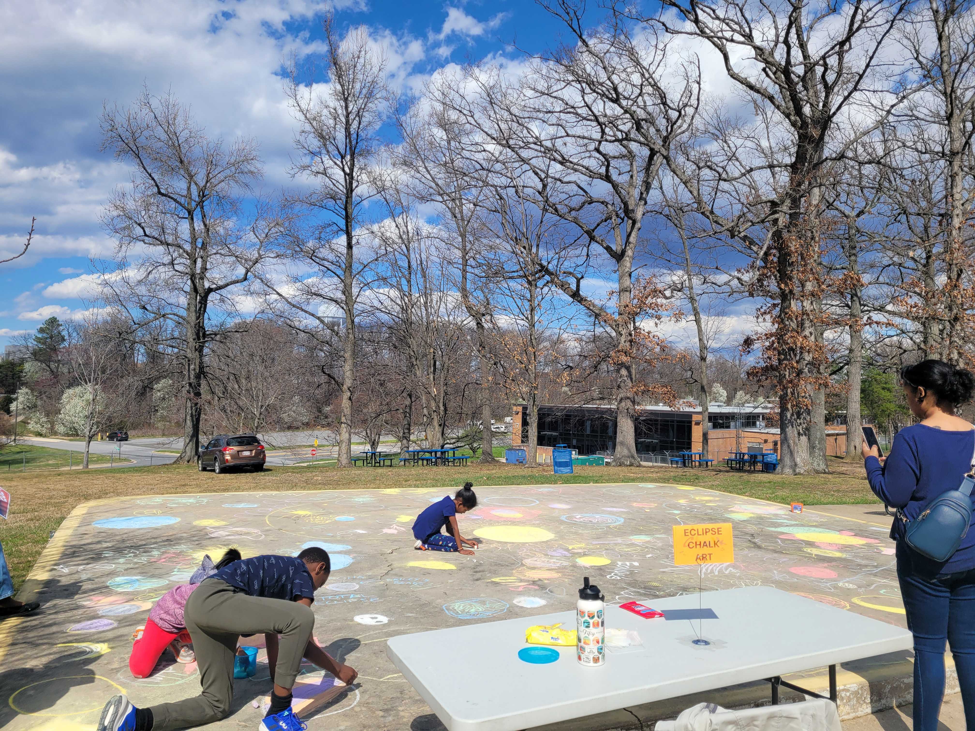 Kids sitting on a concrete pad are using chalk to draw Sun and space-themed drawings on the ground.
