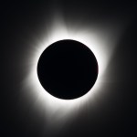 Against a black background is a total solar eclipse. In the middle is a black circle – the Moon. Surrounding it are white streams of wispy light, streaming out into the sky.