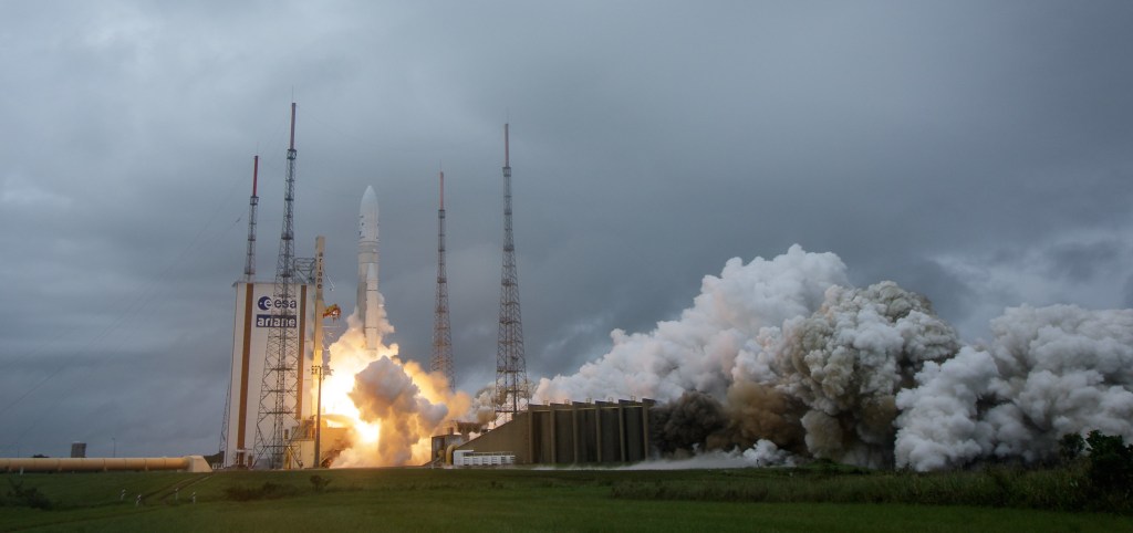 Webb launches on a cloudy day from an Arianne 5 rocket. The image shows the rocket just lifting off the pad with a plume of orange fire and a plume grey blue smoke extending well to the right against a slate blue sky.