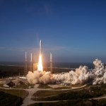 a pulled back view of a Kennedy Space Center launch pad with rocket lifting off against a clear blue sky with smoke plume spreading beyond the pad.