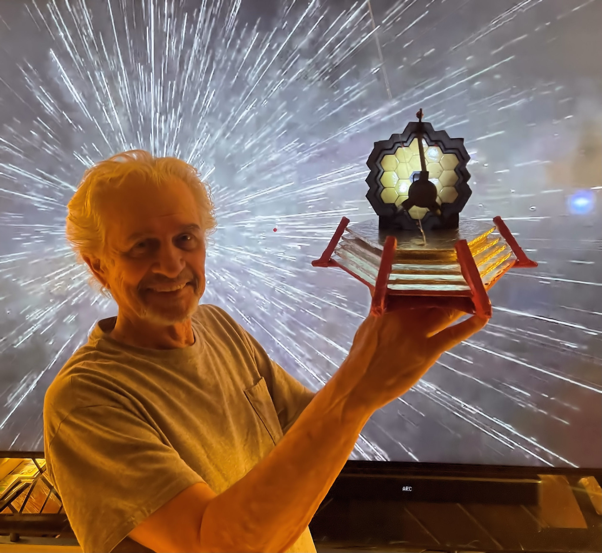An older gentleman holds a model of JWST he built in front a screen radiating light beams.