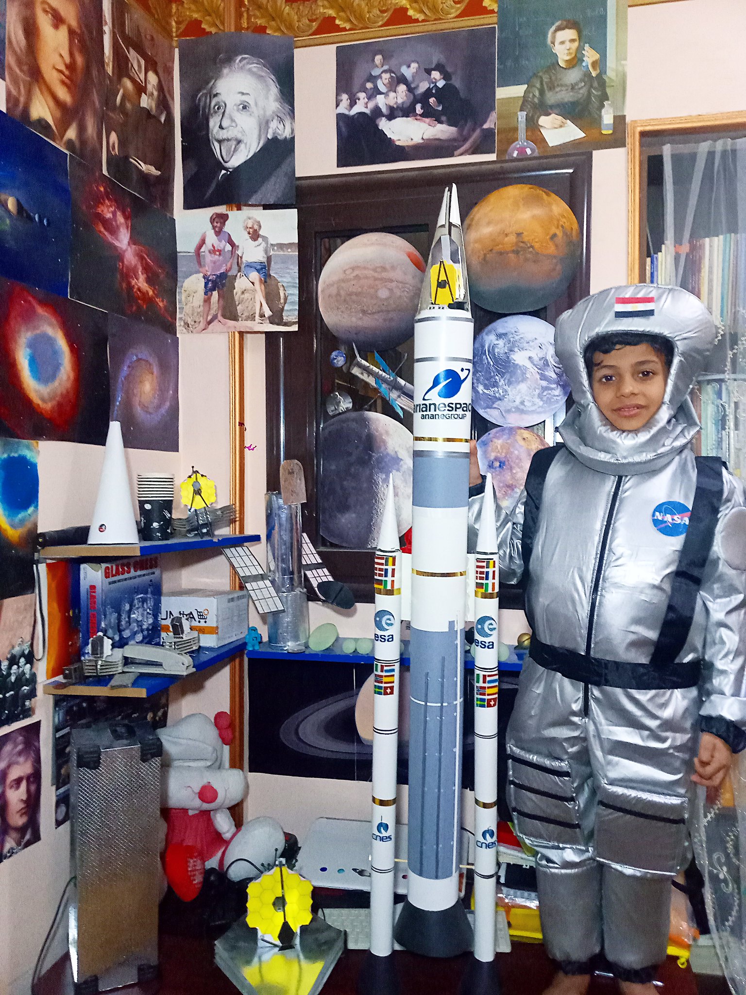 A young boy dressed in a space suit along side a rocket and a number of JWST models he built and surrounded by space images.