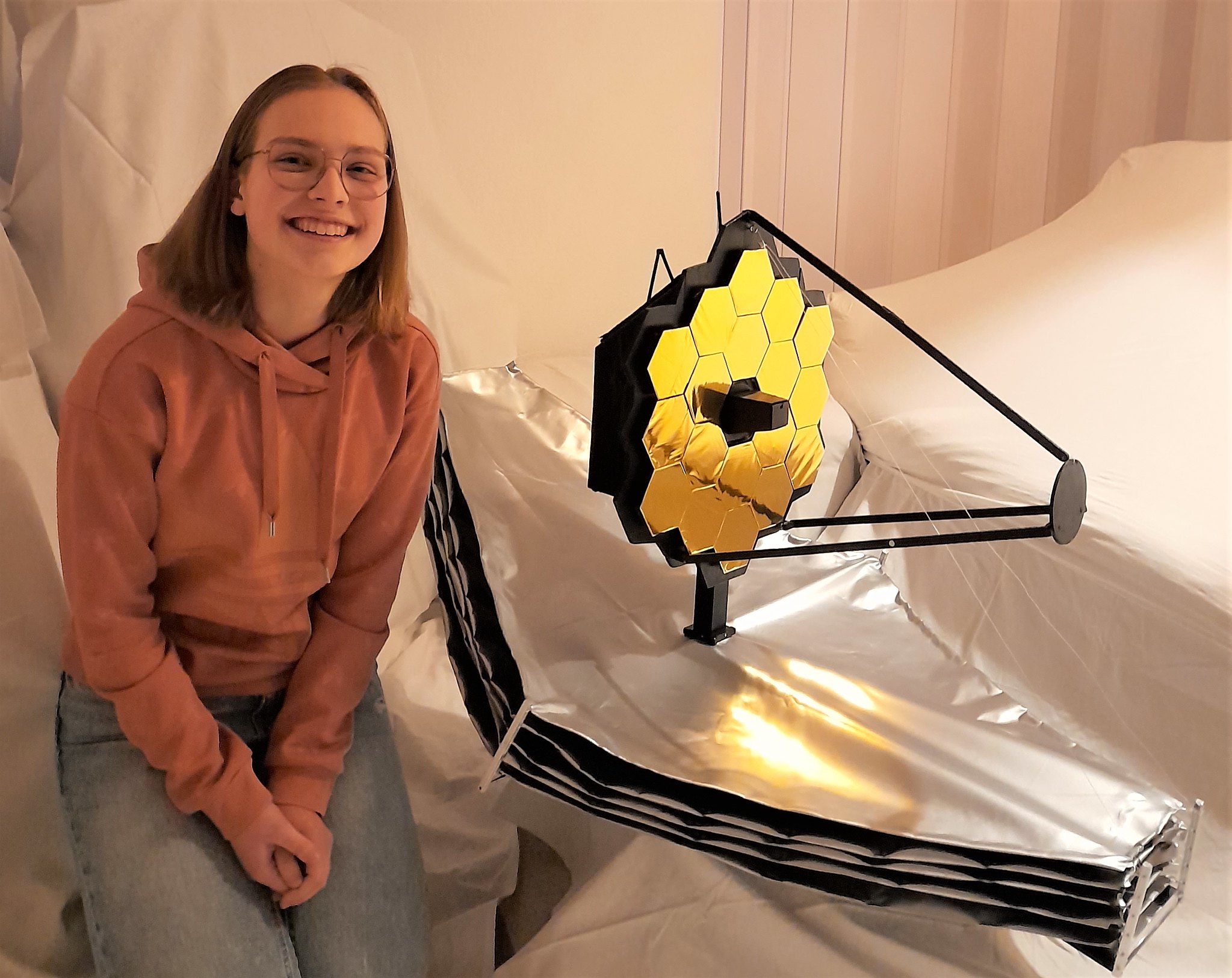 A young lady sits next to a very large scale model of JWST she built.