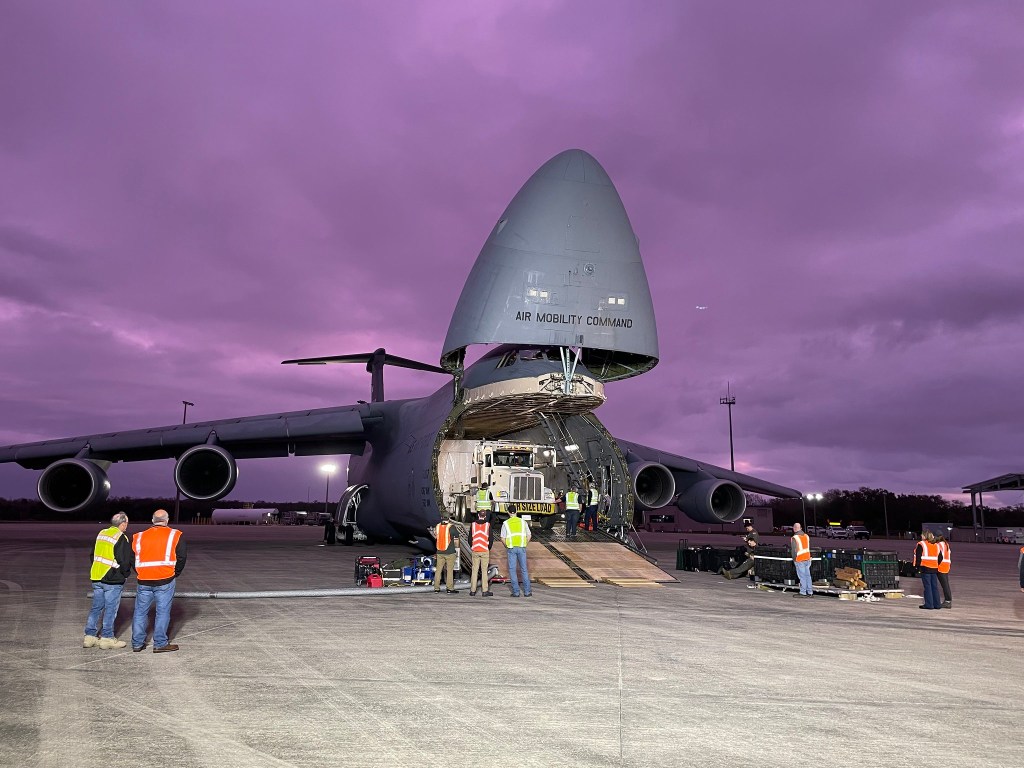 A large cargo plane sits on the runway with the nose cone open to reveal a semi truck inside unloading the GOES U satellite at KSC.