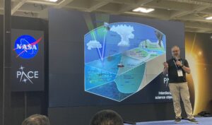 A man wearing glasses, a dark polo shirt, khakis and a nametag on a lanyard stands in front of a large display made of nine screens combined into one. On the screen is a visual showing PACE study areas, labelled on a schematic of the ocean, atmosphere and terrestrial ecosystems.