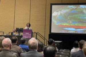 A woman in a light-colored blazer stands behind a podium on a dais, giving a presentation. On the right side of the image, a screen shows a colorful visualization of Earth, with concentrations of different types of phytoplankton in the oceans represented with different colors. In the foreground of the image are the backs of attendees' heads.