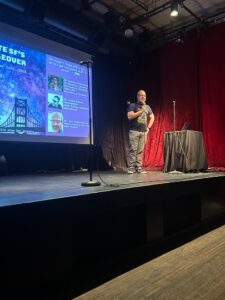 A man stands in the image upon a stage. The black stage takes up a majority of the bottom half of the image. The man holds a microphone and is looking to the right in the image, out to the audience. Behind him to the right is a bright red curtain, and to the left is a projected image of a presentation, which is on a slide that is mostly blue and purple in color.
