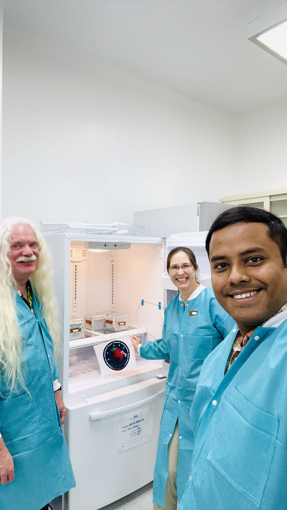 Portrait three people: a Caucasian man with a mustache and long blond-grey hair; a Caucasian woman with glasses and brown hair in a ponytail; and an African American man with short hair. All are wearing blue lab coats and smiling as they prepare their experiment.