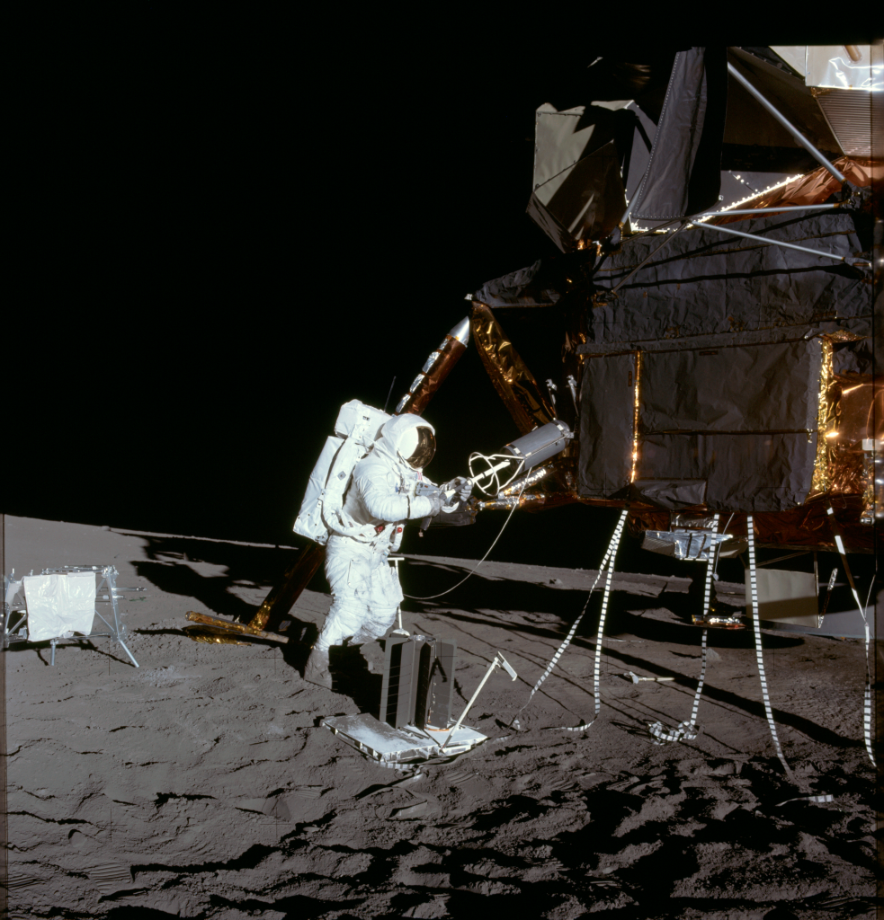 The astronaut is standing on the Moon next to the landing module. He is extracting a round cylinder from a storage compartment on the side.