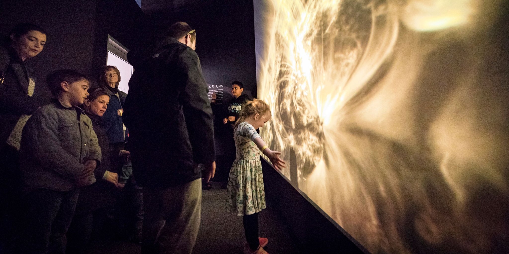 A little girl stands in front of a large projection of the Sun with her arms outstretched. Several other people stand behind her.