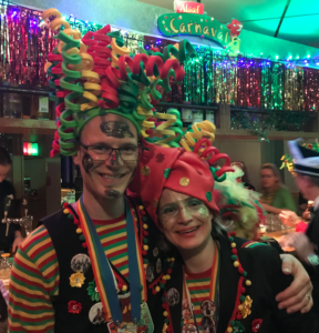 A man and a woman are standing side by side, the man to the left with his arm around the woman's shoulder. They both have glasses on and their faces are covered in a glittery face paint. They wear green, yellow, and red striped shirts and black vests that have colorful puns on them. They both have large hats upon their heads with green, yellow, and red spirals on them. The lighting in the room is dim and there are streamers hanging on the walls behind them.