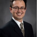 Formal portrait of man with dark hair and glasses in black suit with black and gold striped tie, white shirt