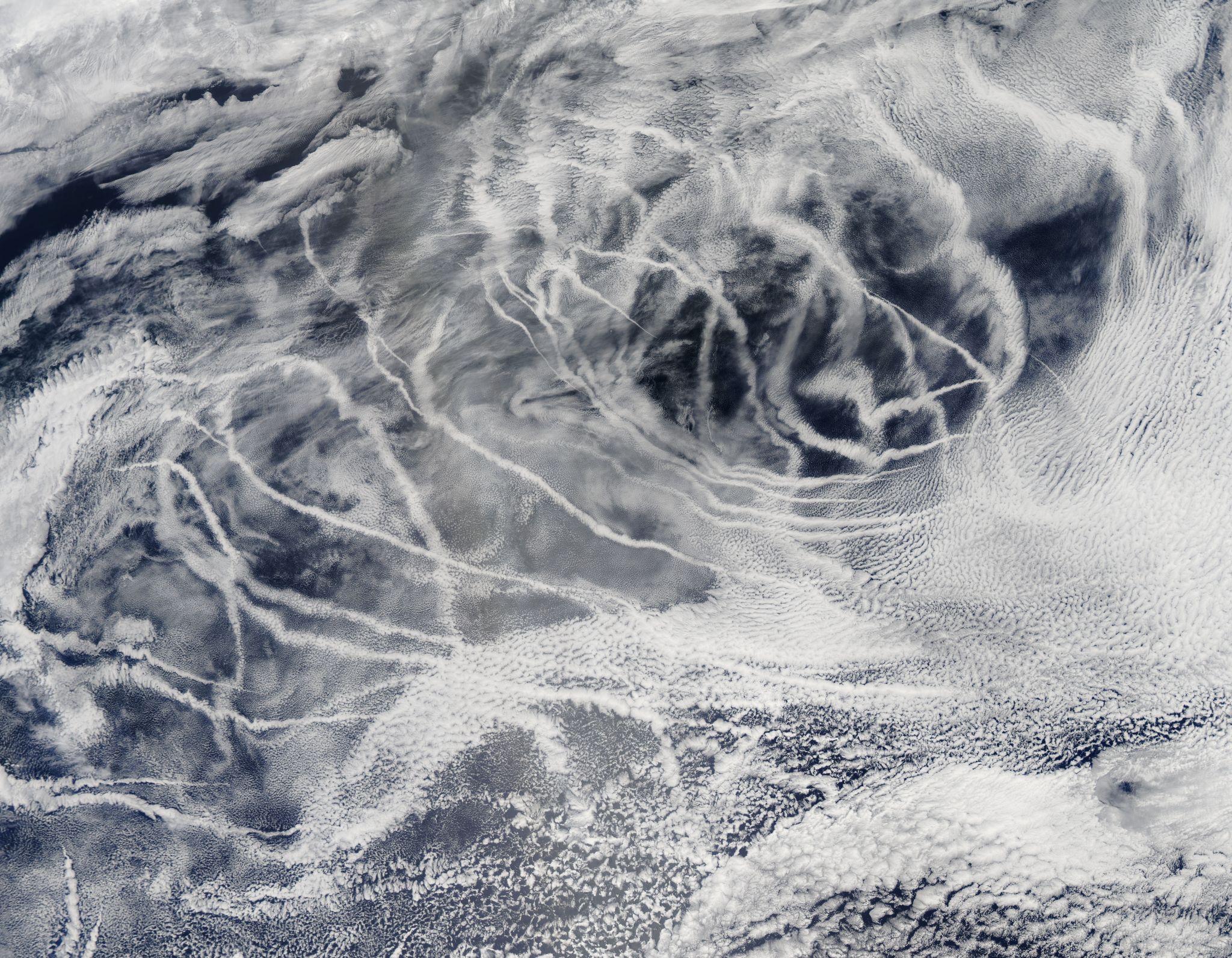 The image is an aerial view overlooking the ocean and clouds. The background of the image is the deep blue of the ocean while the foreground shows white wispy clouds. The clouds cover most of the image, so blue is only peeking through in some spots. The clouds near the borders of the image are thin but cover a lot of area. Near the center of the image are clouds tracing streaks through the sky. The streaked clouds – ship tracks – primarily run diagonally in the direction of bottom right up to top left.