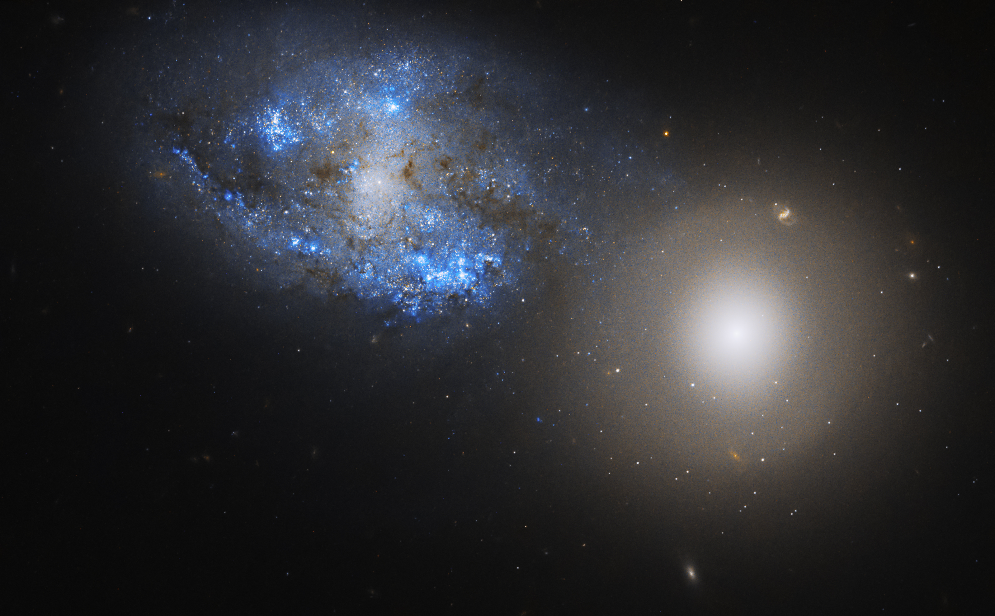 Two side-by-side galaxies fill the image. The leftmost galaxy is a bright spiral galaxy bursting with blue stars. The rightmost is a lenticular galaxy, emitting a hazy glow. Distant stars and galaxies dot the background.