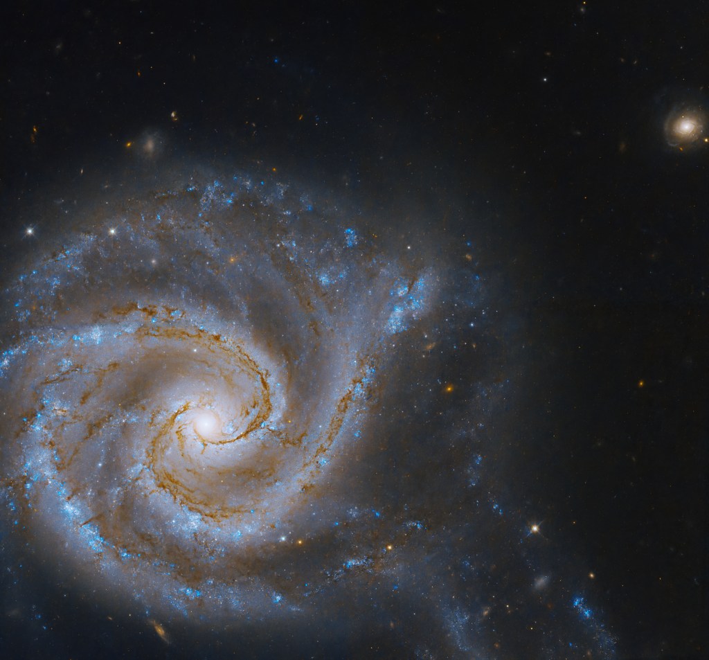 A massive spiral galaxy fills the lower left of the image. Spiral arms full of dark brown dust and bright blue stars extend out from the yellow galactic core, all against black space dotted with more distant galaxies and stars.