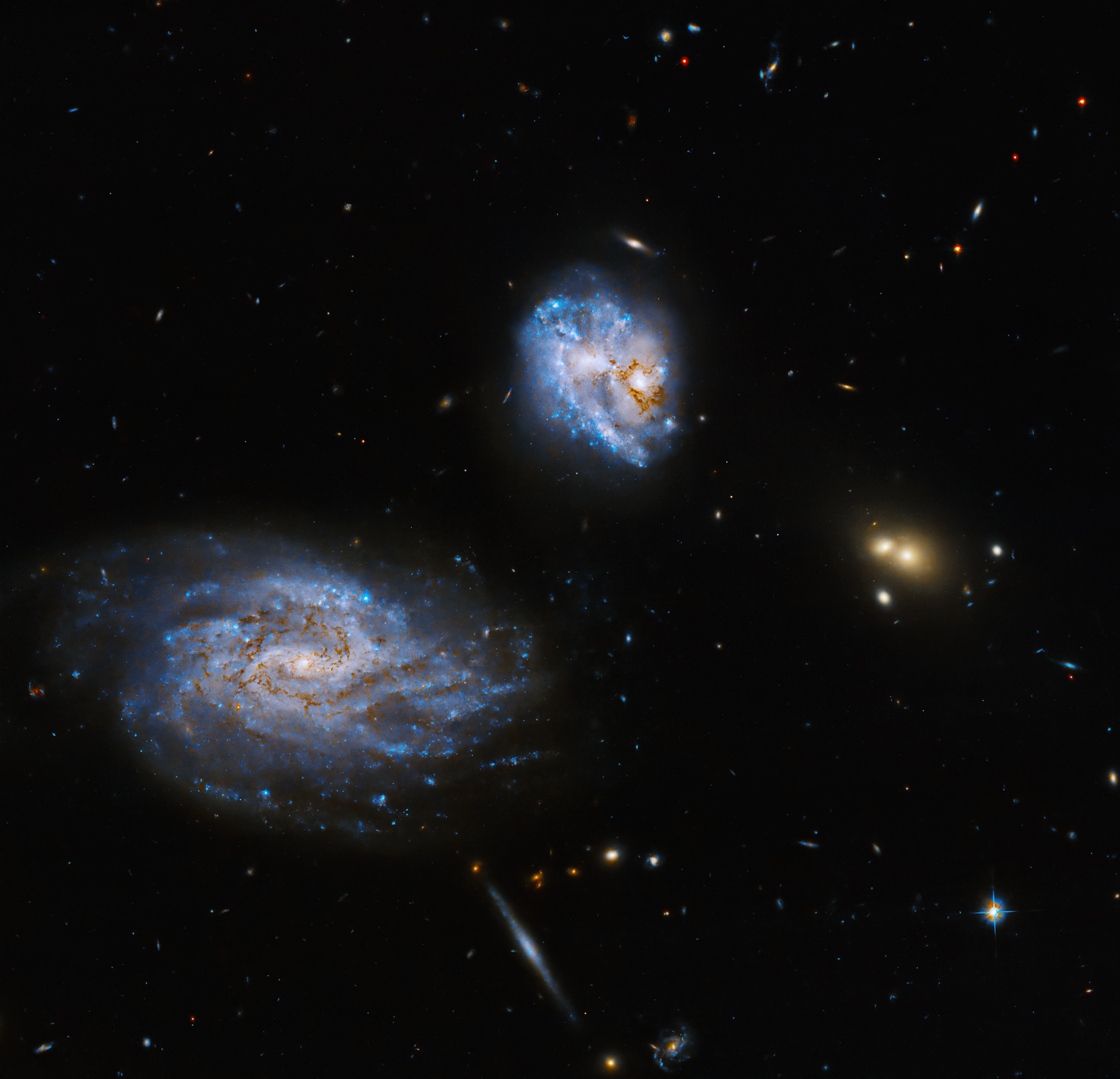 Near the top center of the image, a galaxy full of bright blue stars shines. To its lower left is a spiral galaxy, also full of blue stars and dark brown dust. Other distant galaxies and stars fill the black background of space.