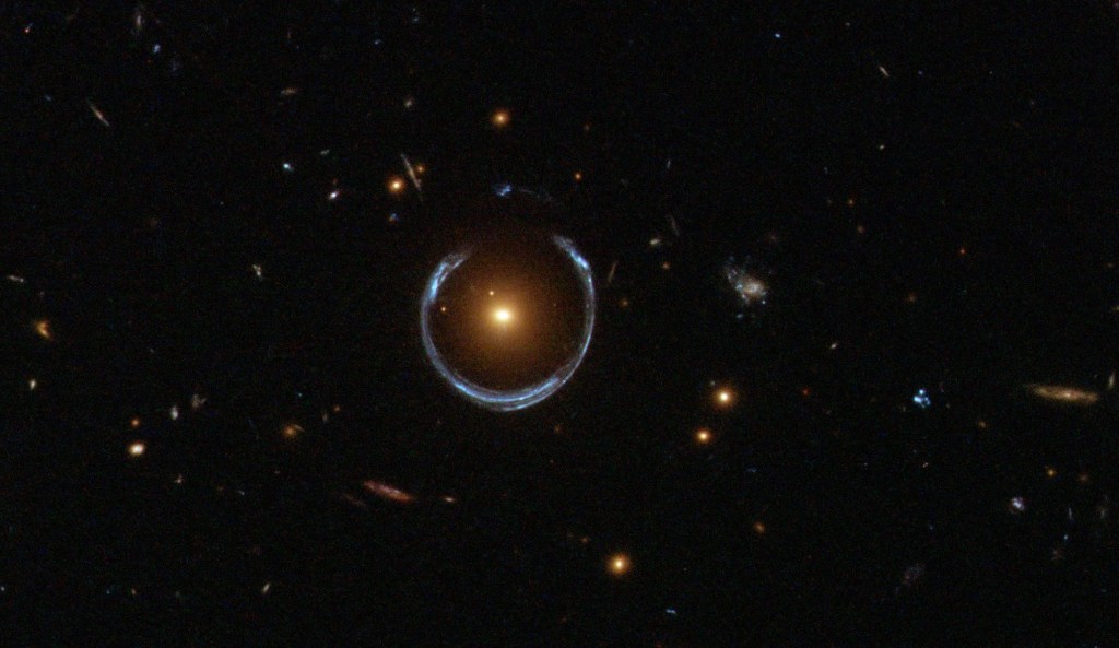 Black background dotted with galaxies. A reddish galaxy at image center with a bluish-white ring curving almost completely around it.