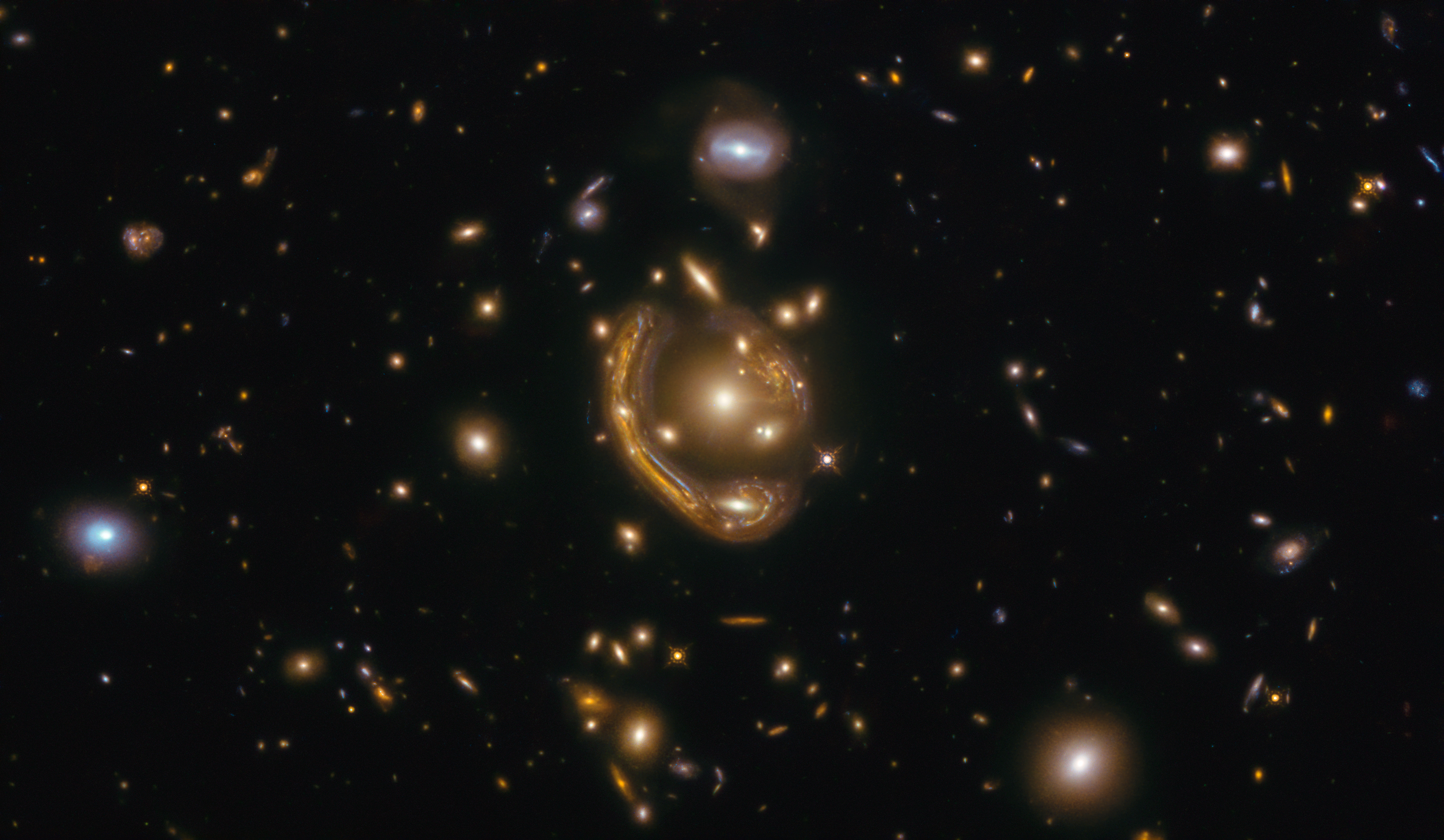 A black background dotted with galaxies. Image center holds a cluster of galaxies with a distinct arc nearly surrounding it.
