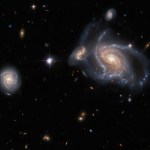 A collection of galaxies. On the right side a large spiral galaxy with swirling, twisted arms is flanked by a smaller, but still detailed, spiral behind its arm on the left, and a smaller spiral above it. On the left side is a fourth, round spiral galaxy seen face-on. Between them lies a single bright star. Several stars and distant galaxies dot the background.