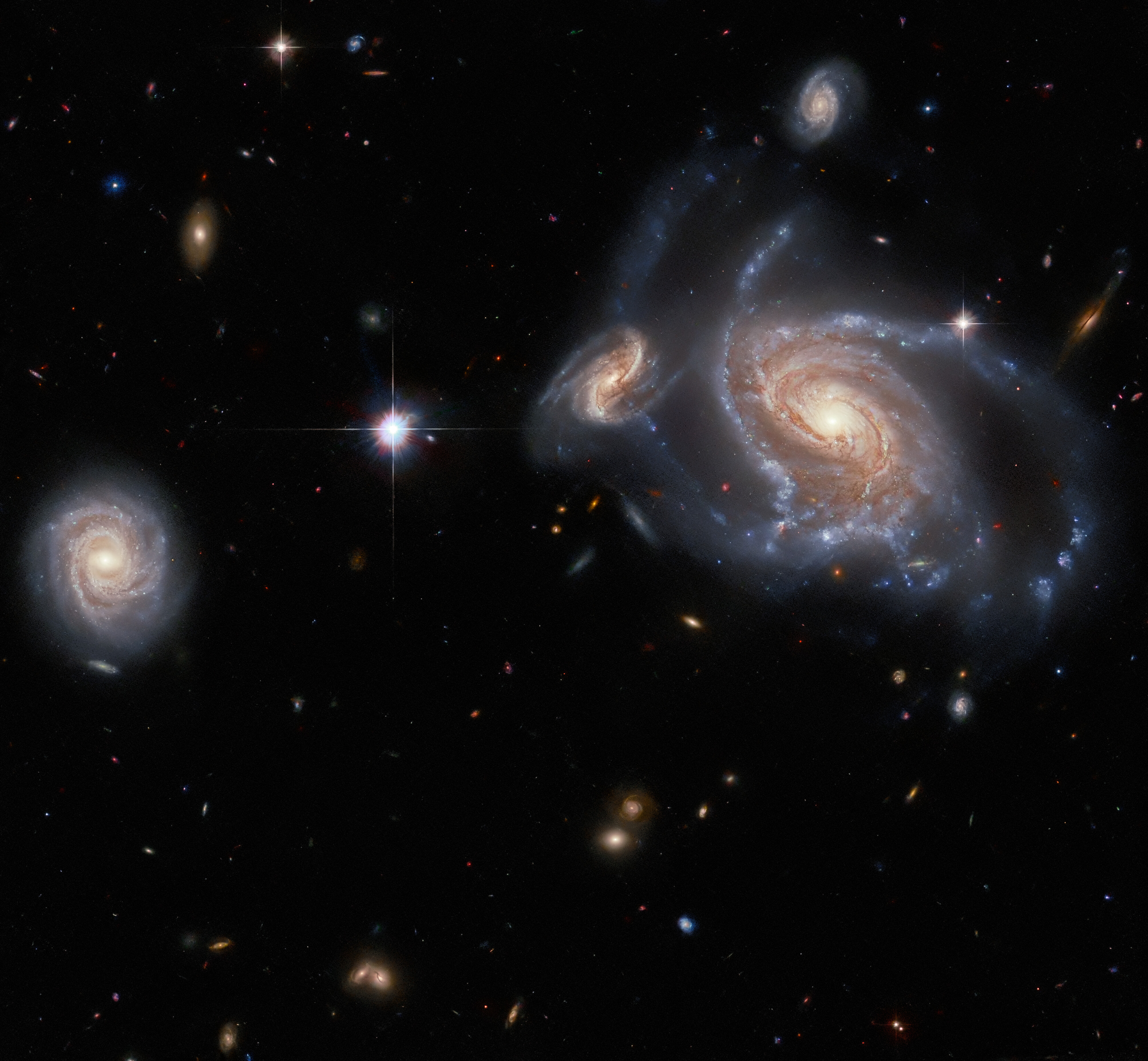 A collection of galaxies. On the right side a large spiral galaxy with swirling, twisted arms is flanked by a smaller, but still detailed, spiral behind its arm on the left, and a smaller spiral above it. On the left side is a fourth, round spiral galaxy seen face-on. Between them lies a single bright star. Several stars and distant galaxies dot the background.