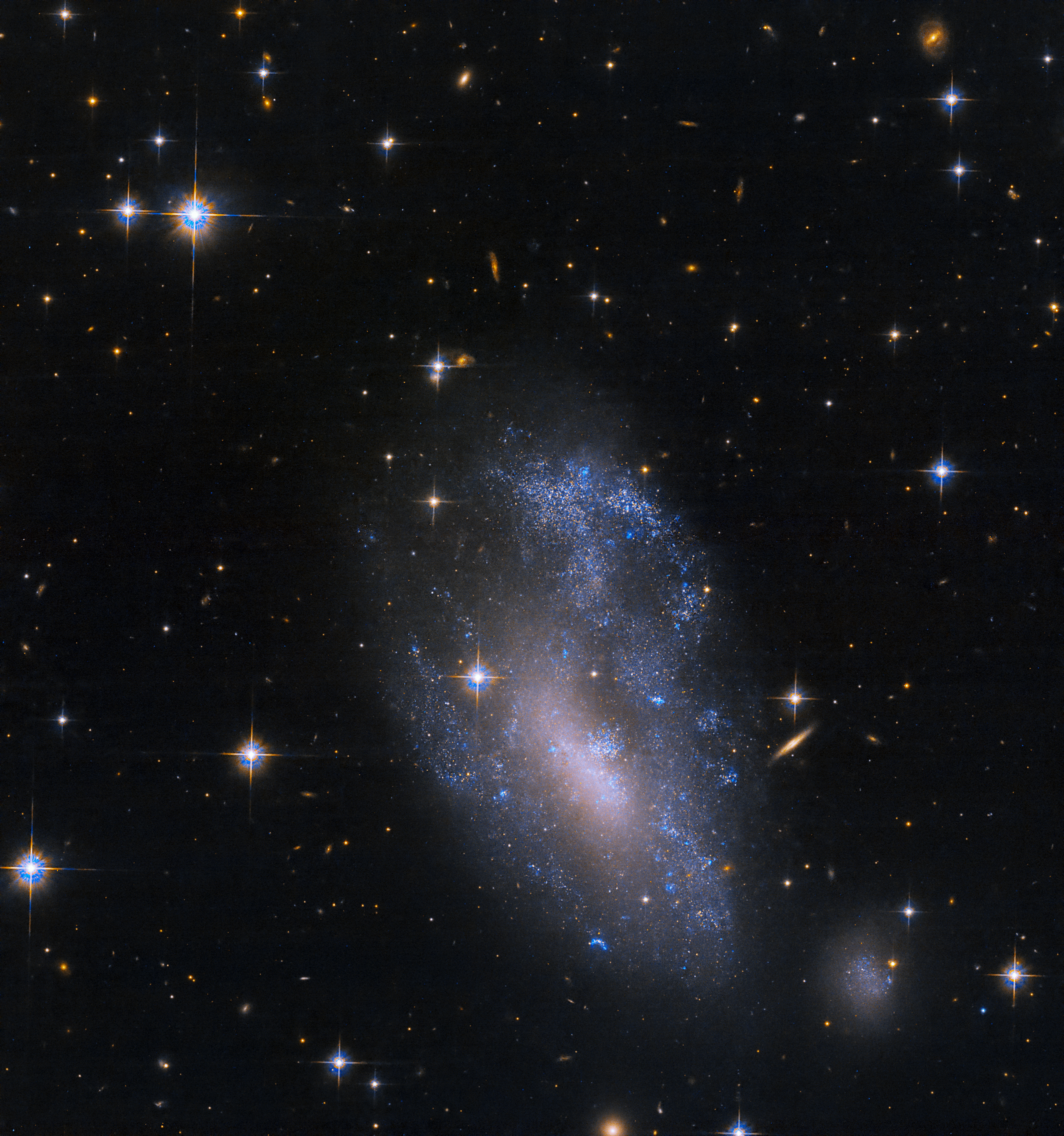 A scattered grouping of blue and violet stars shines near the lower half of the image. Distant galaxies fill the black background of space, and bright stars with diffraction spikes are visible throughout.