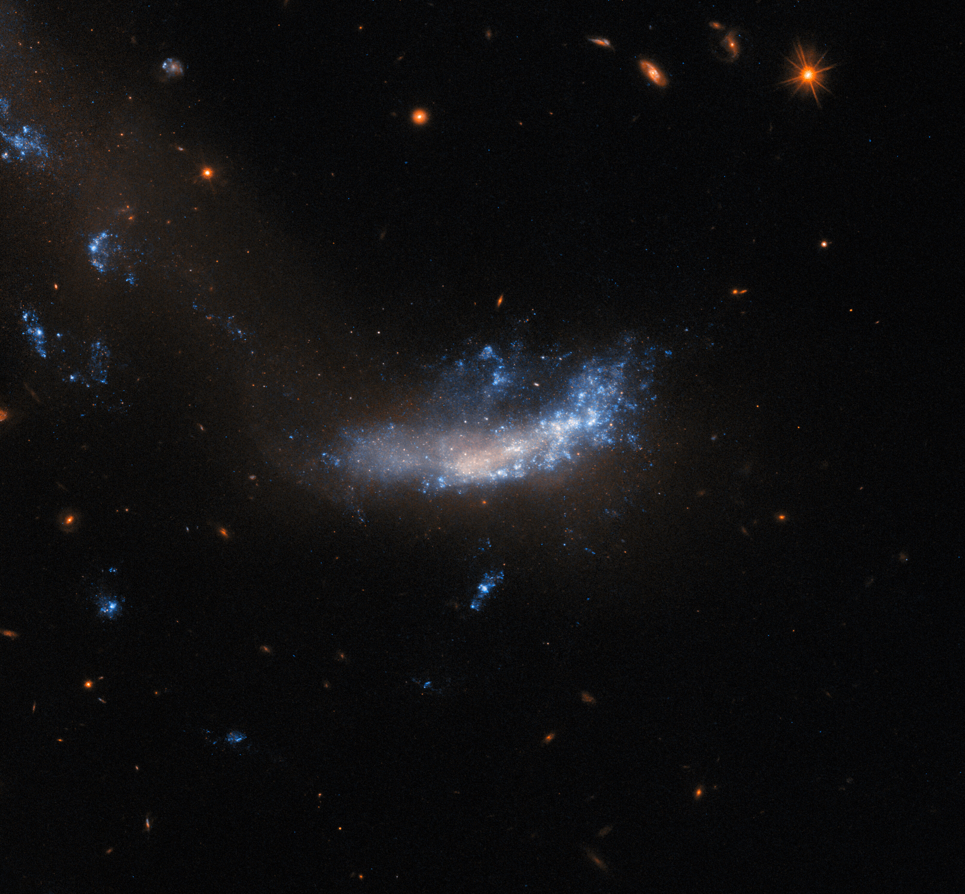 A galaxy that is flat and misshapen. Above and on its right side are plumes of shining gas and dust, while its center and left side are dimmer and patchier. A trail of dark, dim dust stretches from below the galaxy up and off to the left, where there are three more bright patches. The background around the galaxy is quite dark. It holds a few small background galaxies and one bright, foreground star.