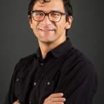 wavy dark-haired man with glasses in black short-sleeved dress shirt in formal headshot