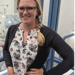 woman with blonde braid and glasses, brown cardigan over elephant-print batik-style blouse, hand on her hip, smiling in front of the Europa Clipper clean room window