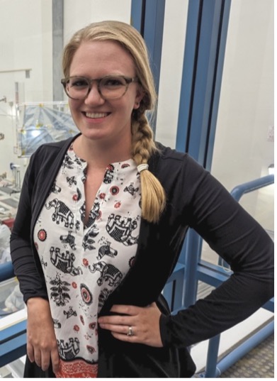 woman with blonde braid and glasses, brown cardigan over elephant-print batik-style blouse, hand on her hip, smiling in front of the Europa Clipper clean room window