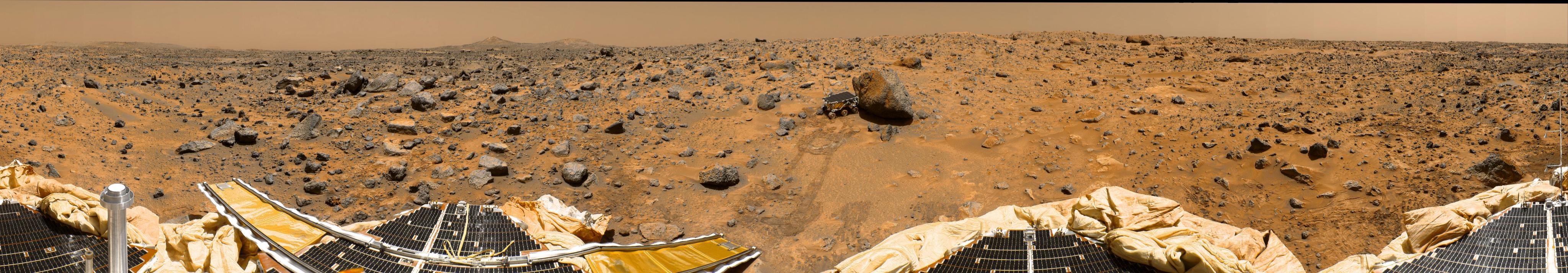 A rover the size of a child's wagon explores a large rock on the surface of Mars. The Pathfinder science station and rover tire tracks are visible in the foreground.